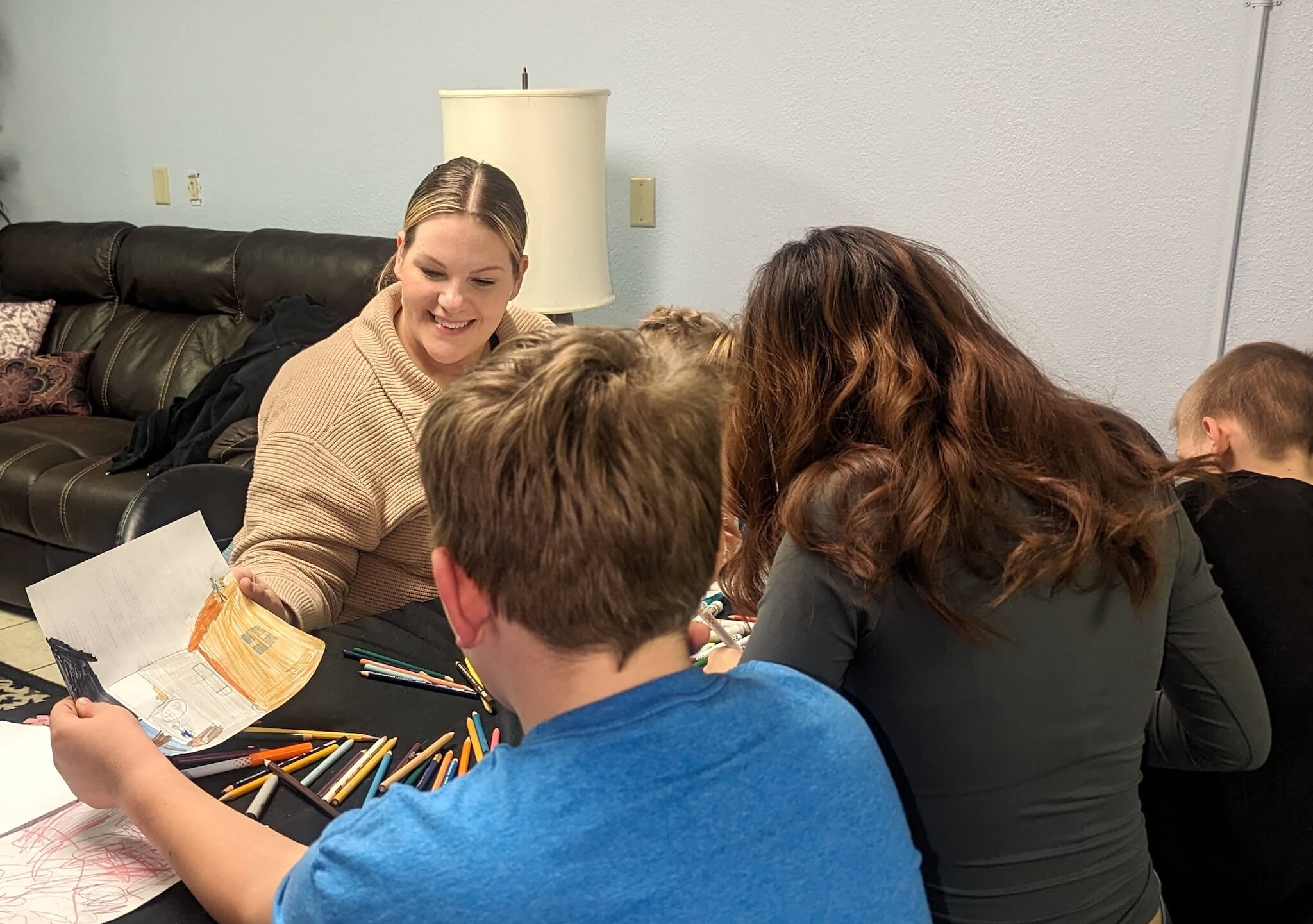 Two adults and two children working at a table with colored pencils and art paper. Two of them are holding and looking at a drawing of several buildings.